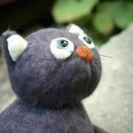 A vintage felt plushie cat in a garden looks with curiosity at something above, macro photogr...webp
